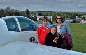 Flugplatz Ottenschlag02, © Union Segelfliegergruppe Ottenschlag Drei Personen stehen neben einem kleinen Flugzeug auf einem Flugplatz mit grüner Wiese und bewölktem Himmel im Hintergrund.