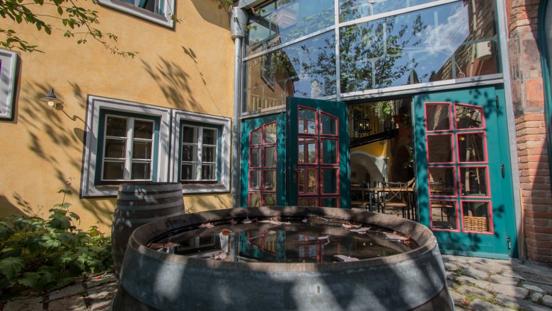 img_0063, © Michael Holzmayer Ein Innenhof mit gelber Fassade, grünen Türen und einem Holzfass mit Wasser. Schatten von Bäumen fallen auf die Wand.