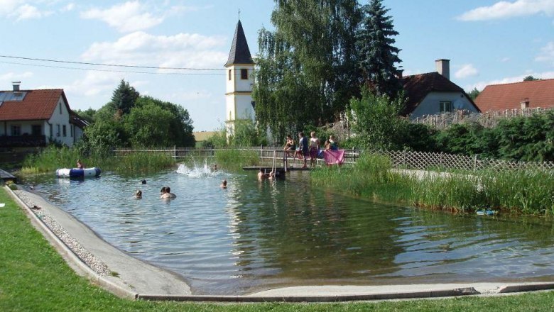 badeteich_obergrünbach, © Gemeinde Lichtenau Ein natürlicher Badeteich mit Menschen, die schwimmen. Im Hintergrund sind ein Kirchturm und Häuser zu sehen.