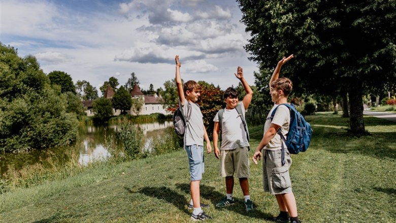 Waldschule Waldviertel, © Waldviertel Tourismus, Matthias Streibel Drei Jungen mit Rucksäcken stehen auf einer Wiese neben einem Teich und heben die Arme in die Luft.