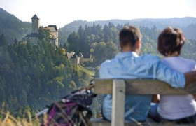 Jugendlagerplatz Rappottenstein, © Waldviertel Tourismus Ein Paar sitzt auf einer Bank mit Blick auf eine Burg in einer bewaldeten Landschaft.