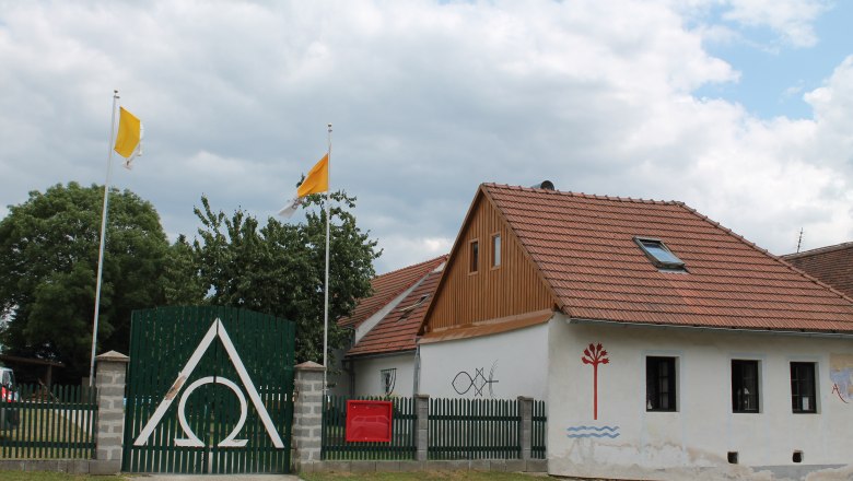 House Agnes 3, © Hannes Mesmann A house with red roof tiles, a green fence with the Alpha Omega symbol, two yellow flags and murals.