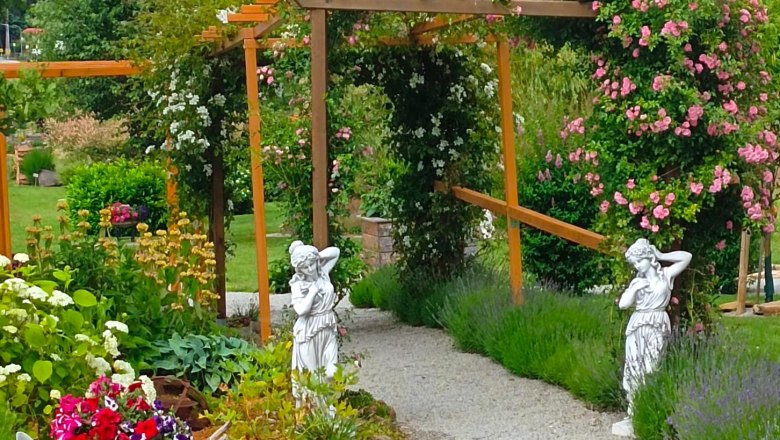 Garten der 100 Rosen_Scherzer, © Garten der 100 Rosen, B.Scherzer Ein gepflegter Garten mit Rosenbogen, Statuen und blühenden Pflanzen.