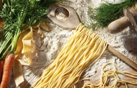 Köstliche Nudeln, © Zimmermanns Teigwaren Frische Nudeln auf einem Holzlöffel mit Mehl, Karotten und Kräutern auf einem Tisch.