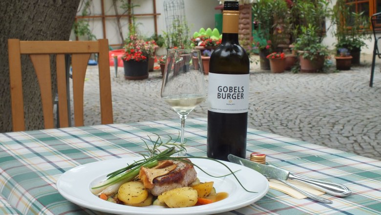 Cobane farm, © Cobaneshof A plate of meat and potatoes on an outdoor table, with a bottle of wine and a glass next to it.