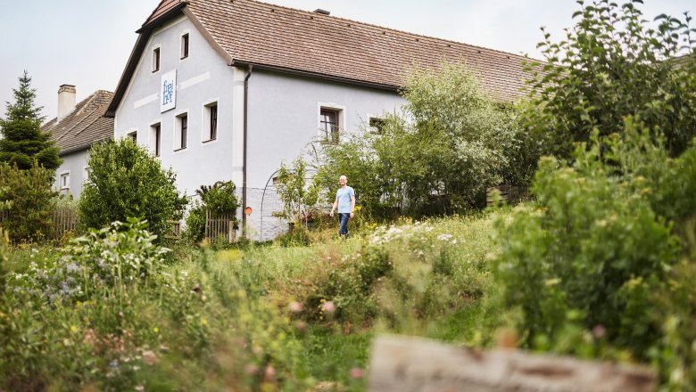 SONNENTOR Freihof, © Waldviertel Tourismus, Gerhard Wasserbauer Ein Mann steht vor einem traditionellen österreichischen Haus, umgeben von üppigem Grün und einem Garten.