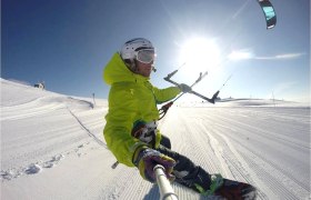 Snowkitekurse, © Snowkitekurse.at Person beim Snowkiten auf einer verschneiten Piste mit blauem Himmel und Sonnenschein.