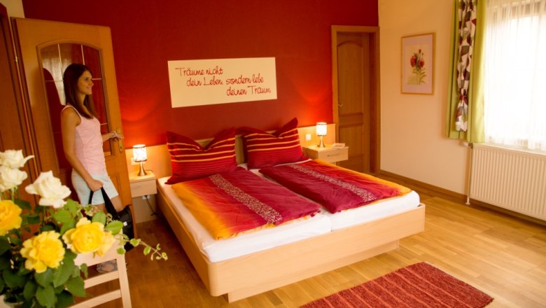Room Danglhof, © Biobauernhof Dangl, Alexandra A cozy bedroom with a red wall, double bed and flowers. A woman enters the room.