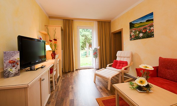 Mohnblütenzimmer, © Johann Frank Gemütliches Zimmer mit roten Akzenten, Fernseher, Sofa, Sessel und Blick auf den Garten durch eine Glastür.