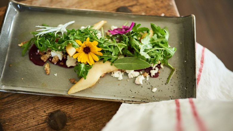 Bio-Gasthaus Leibspeis', © SONNENTOR/ @nudlholz.at Ein kunstvoll angerichteter Salat mit Rucola, gelber Blume und Birnenscheiben auf einem rechteckigen Teller.