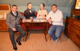 Museum Neupölla, © Gemeinde Neupölla Zwei Männer sitzen an einem Tisch mit zwei Pappfiguren in einem Raum mit antiken Möbeln.