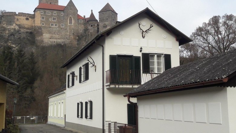Apartments Zum Burgblick, © Jürgen Mokesch A white house with green shutters and deer antlers on the wall, with a castle on a hill in the background.