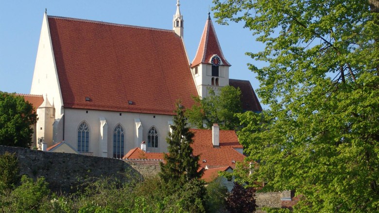 Pfarrkirche St. Stephan, © Stadt Eggenburg Pfarrkirche St. Stephan mit rotem Dach und Turm, umgeben von Bäumen und einer Steinmauer.