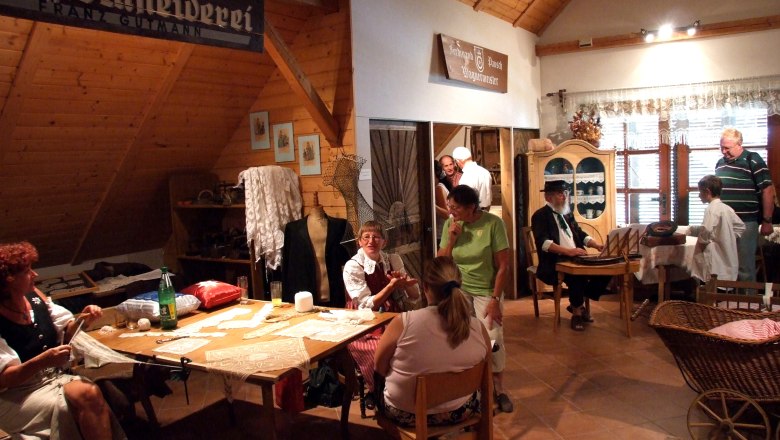 Heimatmuseum Kautzen, © Heimatmuseum Kautzen Innenansicht eines Heimatmuseums mit Menschen in traditioneller Kleidung, die handwerkliche Arbeiten zeigen.