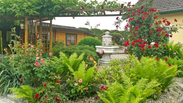 Garten der 100 Rosen__B.Scherzer, © Garten der 100 Rosen, B.Scherzer Ein gepflegter Garten mit Rosen, Farnen und einem kleinen Brunnen, umgeben von einem Holzhäuschen und einer Pergola.