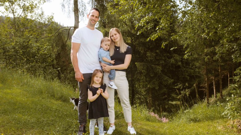 Familie Hahn, © Katja Koppensteiner Fotografie Eine Familie steht auf einer Wiese vor einem Wald. Ein Mann, eine Frau, ein kleines Kind auf dem Arm der Frau und ein weiteres Kind stehen zusammen.
