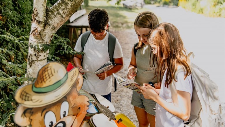 Kittenberger Erlebnisgärten, © Matthias Streibel Drei Kinder mit Rucksäcken lesen Karten neben einer Bärenfigur im Freien.