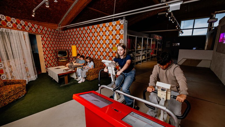 Zurück in der Zeit, © Waldviertel Tourismus, Matthias Streibel Jugendliche auf Fahrradergometern in einem Raum mit Retro-Wohnzimmer-Dekor.