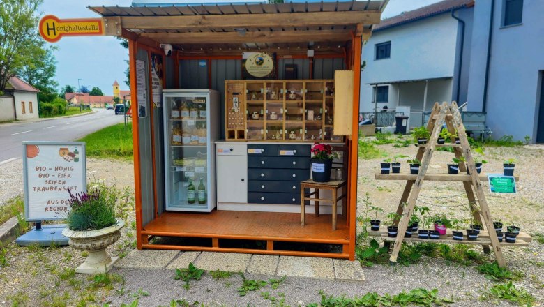 Die Honighaltestelle, © Imkerei Pell Ein kleiner Verkaufsstand am Straßenrand mit Bio-Produkten wie Honig, Eiern und Seifen. Daneben stehen Pflanzen auf einem Holzgestell.