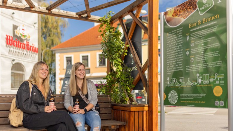 Weitraer Biertour, © Benjamin Wald Zwei Frauen sitzen auf einer Bank vor einem Schild mit Informationen über Bierbrauen, im Hintergrund ein Gebäude mit der Aufschrift 'Weitra Bräu'.