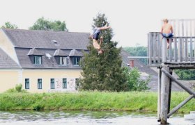 Holzmühlteich, © Ponyhof Holzmühle Ein Junge springt kopfüber von einem Holzsteg in einen Teich, während ein anderer Junge zuschaut. Im Hintergrund ist ein Gebäude mit der Aufschrift 'Gasthaus' zu sehen.
