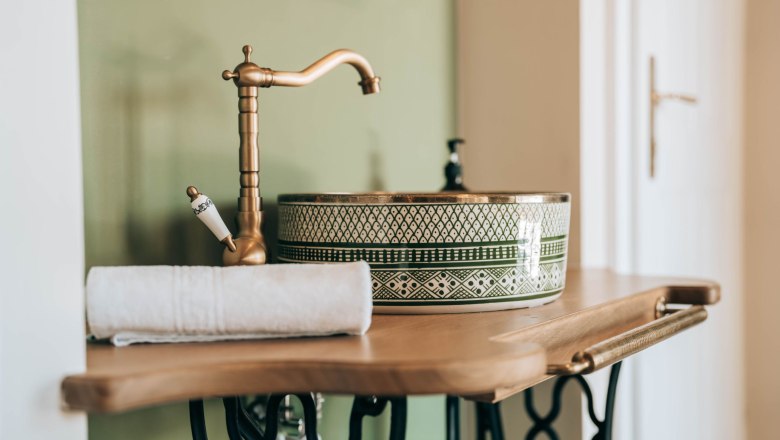 Zum Goldenen Löwen, © Zum Goldenen Löwen Nahaufnahme eines stilvollen Waschbeckens mit goldener Armatur und grünem Muster auf einem Holztisch.