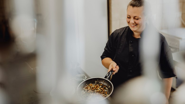 Takes care of the kitchen: Jessica Mann, © Niederösterreich Werbung/Sophie Menegaldo A cook in a black uniform holds a pan of roasted vegetables and smiles.