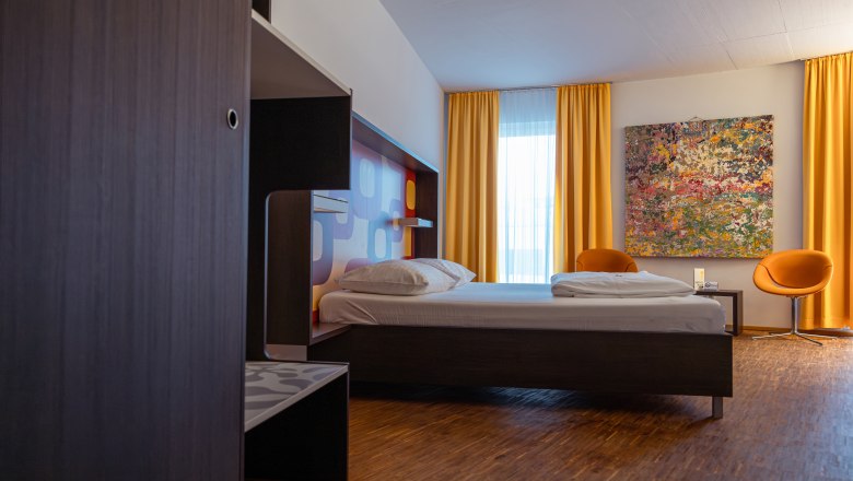 arte Hotel Krems, © Niederösterreich Werbung / Maximilian Pawlikowsky Modernes Hotelzimmer mit Bett, gelben Vorhängen und buntem Gemälde an der Wand.