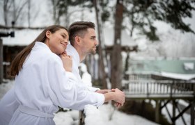 Ein Paar in weißen Bademänteln steht im Schnee und blickt in die Ferne.