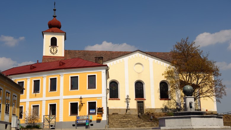 Kirche mit gelber Fassade und Turm mit rotem Dach auf einem Hauptplatz.