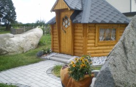 Eine kleine, hölzerne Grillhütte mit grauem Dach, umgeben von Steinen und Pflanzen.