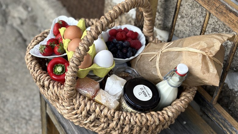 Ein geflochtener Korb mit Eiern, Beeren, Paprika, Brot, Milch und Marmelade auf einer Holzbank.