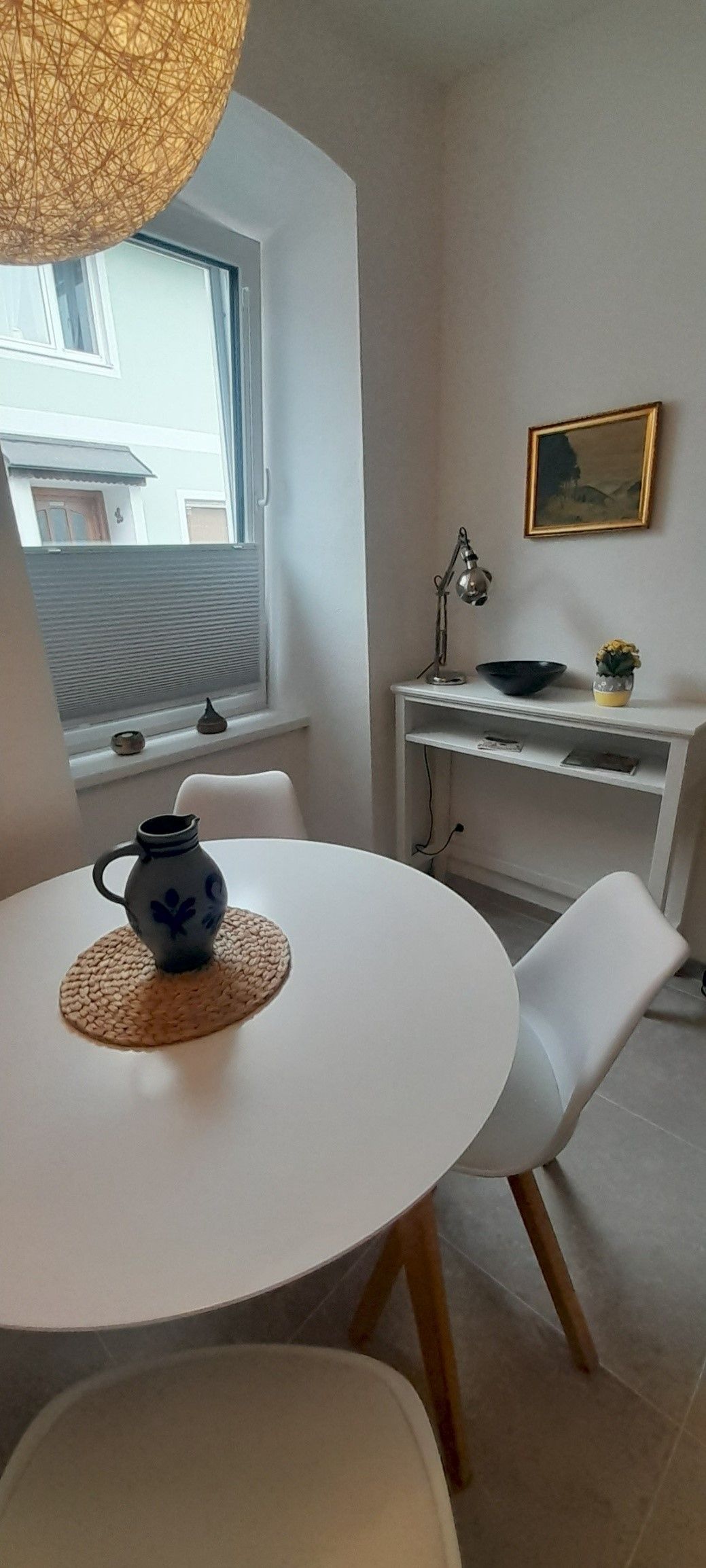 Ein kleiner Essbereich mit einem runden weißen Tisch, zwei weißen Stühlen und einer dekorativen Vase. Im Hintergrund ein Fenster und ein kleiner Tisch mit Lampe.