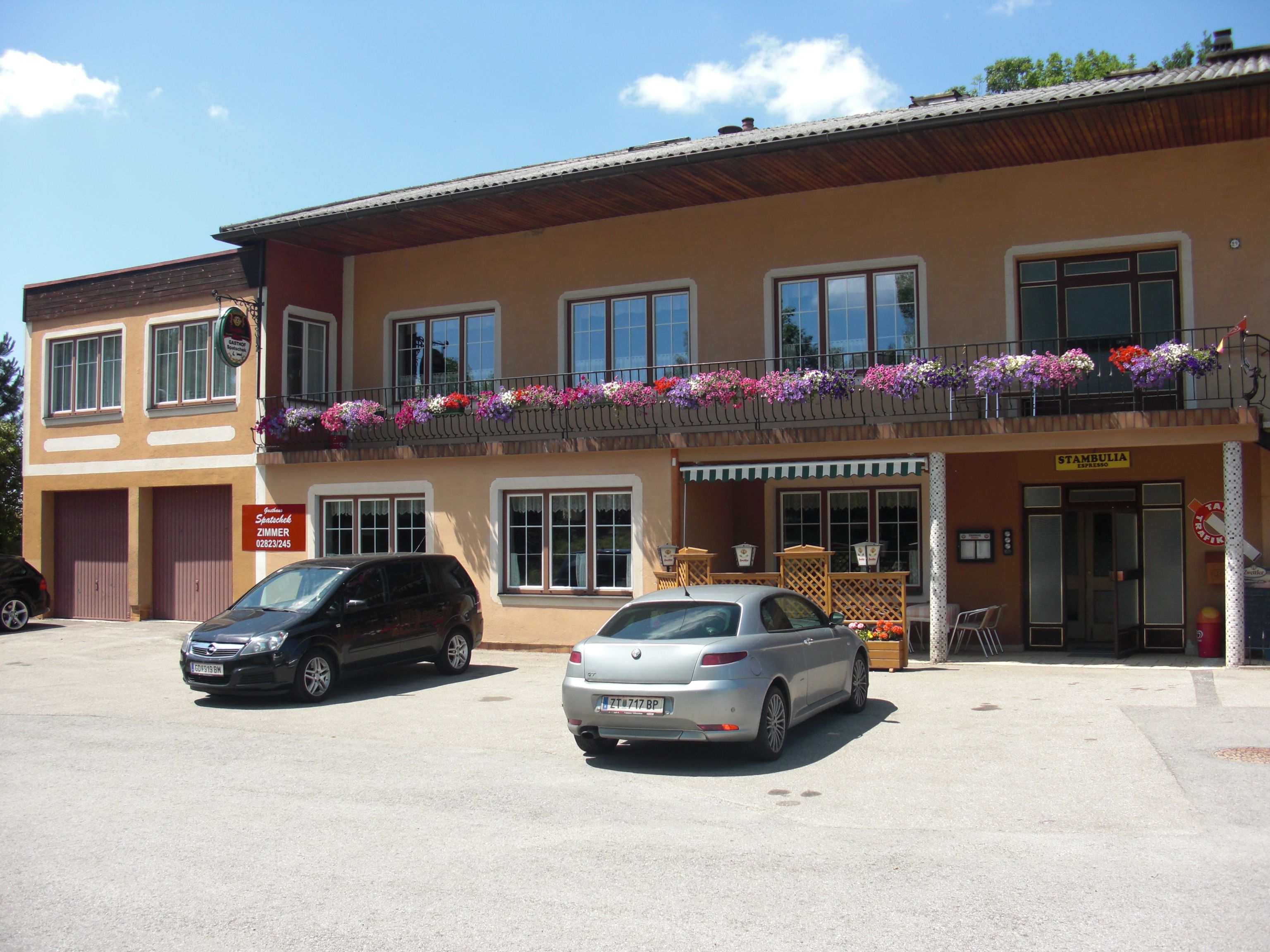 Gasthaus mit Balkon und Blumen, zwei geparkte Autos davor.