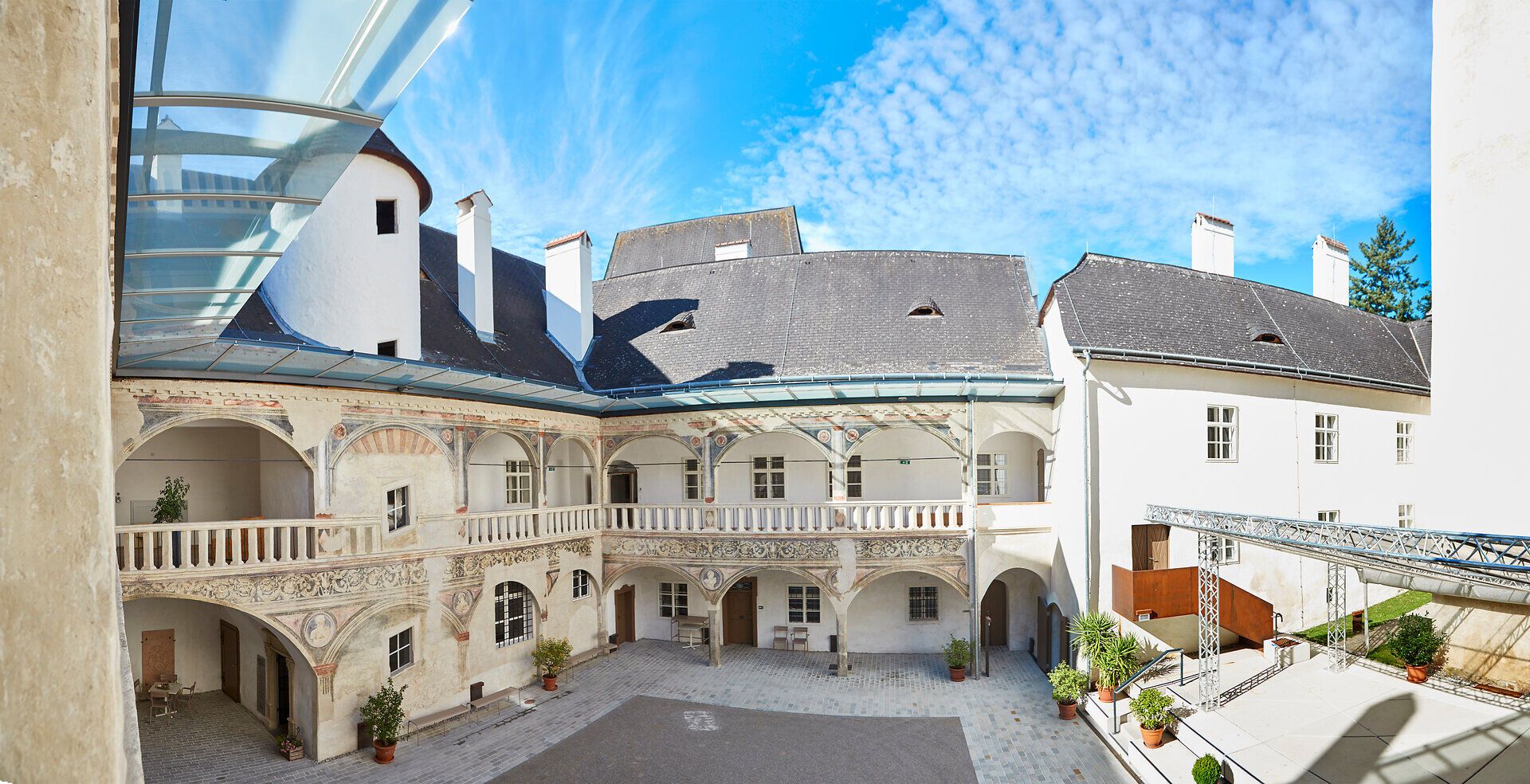 Im malerischen Innenhof des Schlosses Pöggstall entfaltet sich eine harmonische Symbiose aus historischer Architektur und natürlicher Schönheit. Die sanften Wolken am blauen Himmel spiegeln die friedliche Atmosphäre wider, während die kunstvollen Fresken an den Wänden Geschichten aus vergangenen Zeiten erzählen.