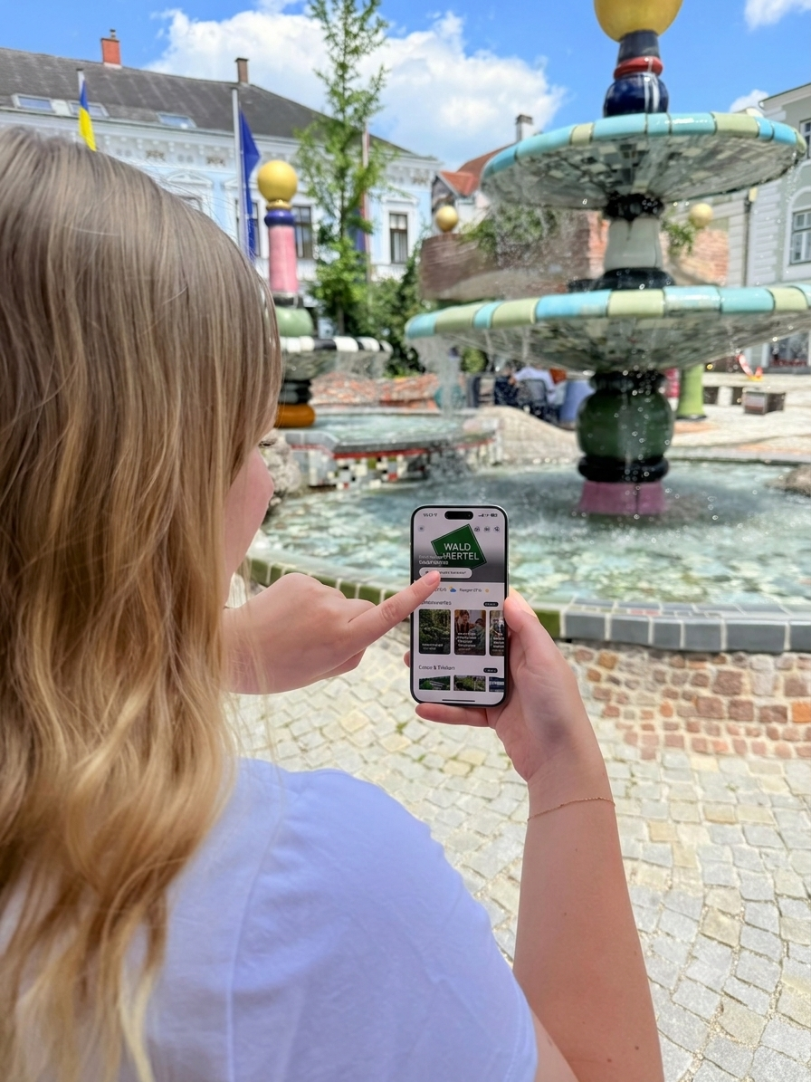 Eine Frau mit einem Handy in der Hand, auf der die Seit "Waldviertel Guide" geöffnet ist. Dahinter ein Stadtplatz mit Brunnen. 