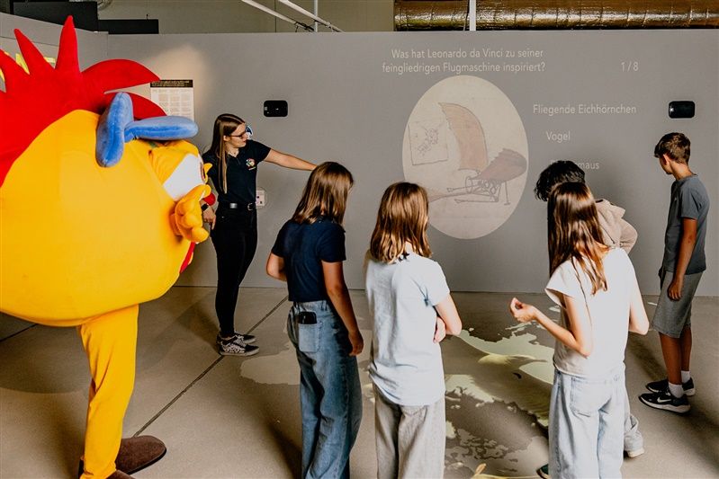 Kindergruppe in einem Museum mit einer Person im Kostüm und einer interaktiven Wandprojektion.