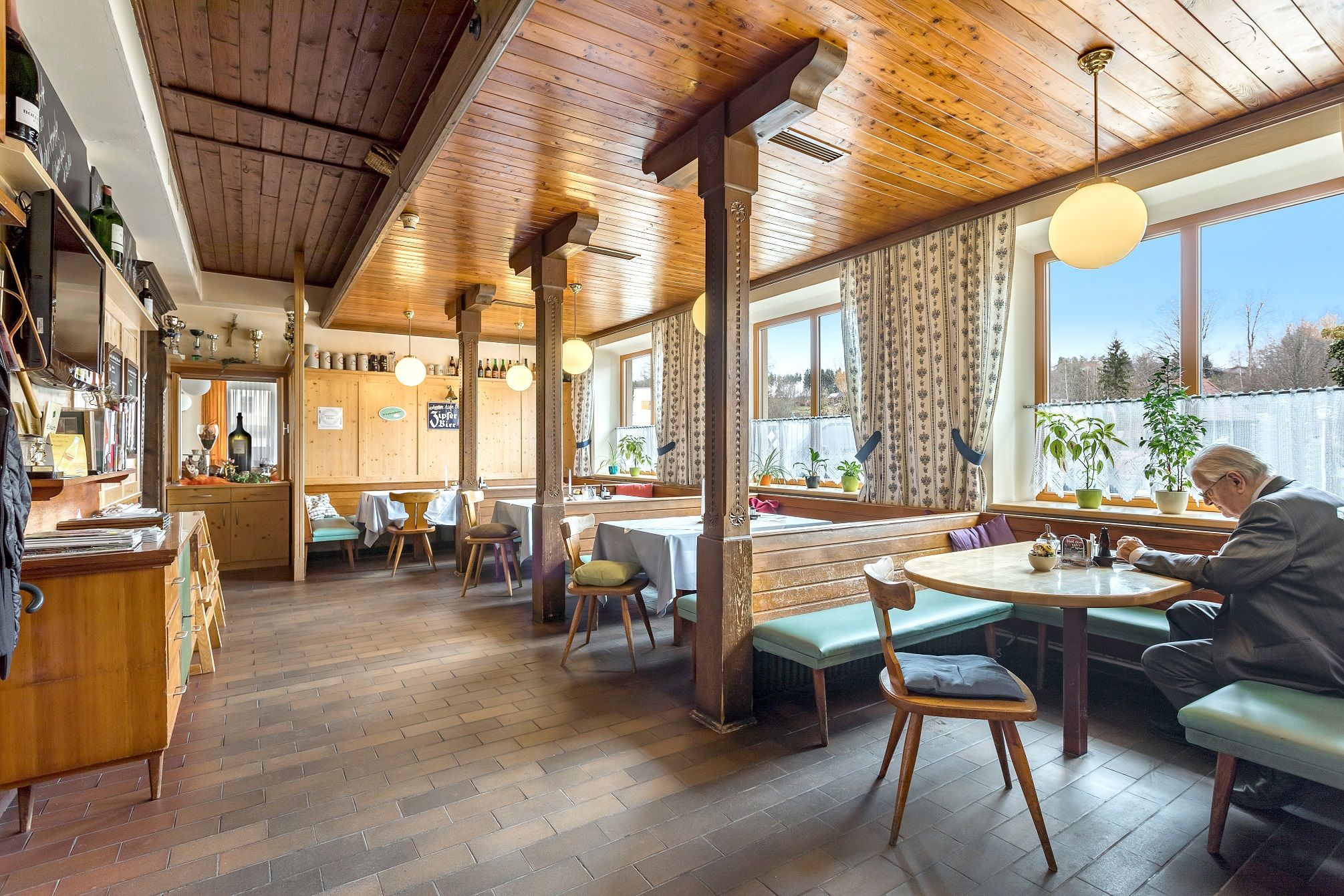Gemütliche Gaststube mit Holzmöbeln und großen Fenstern, ein Mann sitzt an einem Tisch.