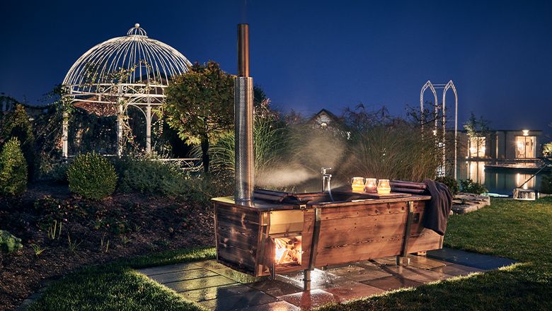 Eine Holzbadewanne mit Feuerheizung im Freien, umgeben von Garten und Pavillon bei Nacht.