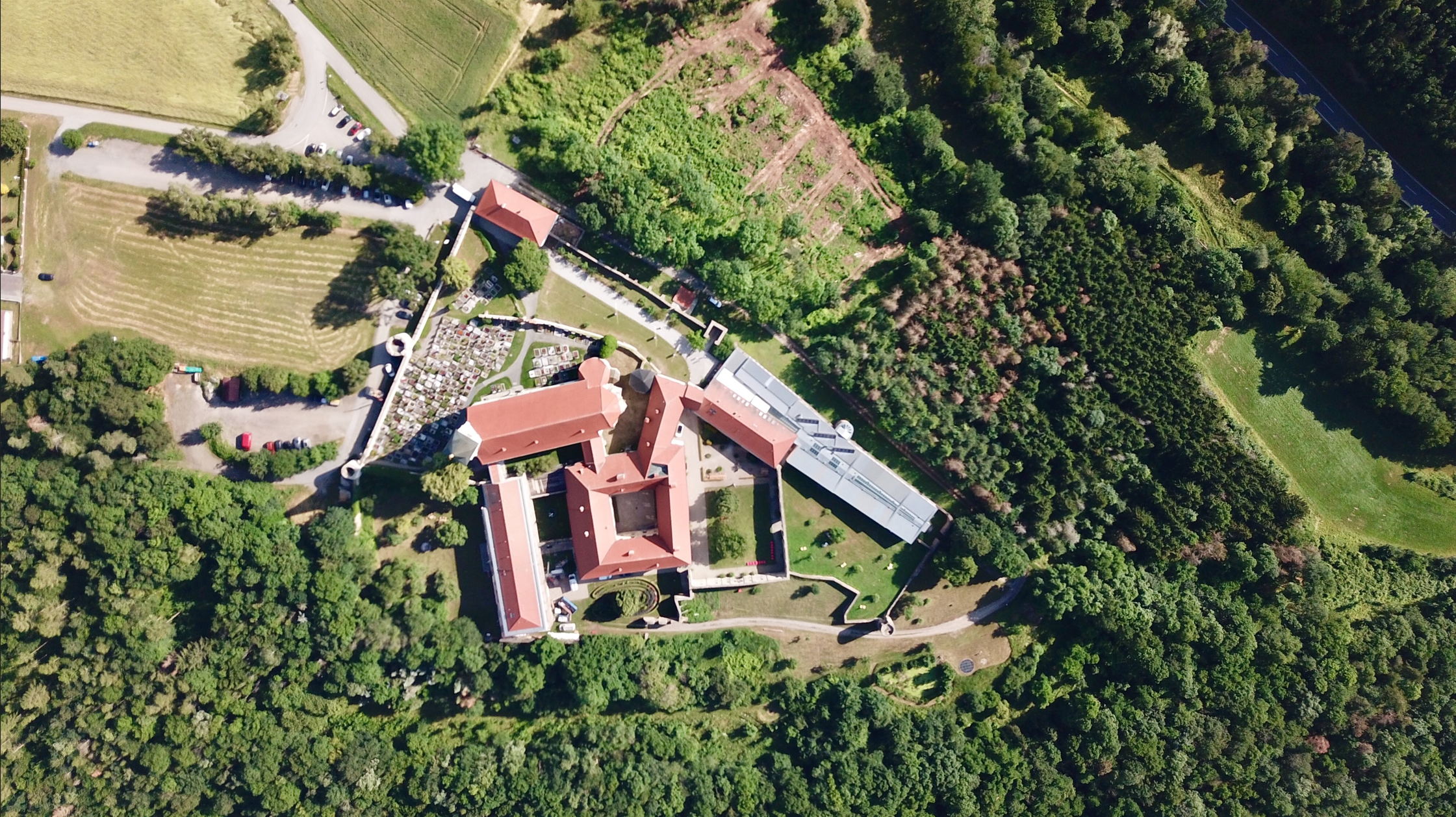 Luftaufnahme eines Klosters mit roten Dächern, umgeben von Wäldern und Feldern.
