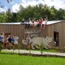 Eine Gruppe von Kindern und Erwachsenen sitzt auf einem Holzgebäude mit der Aufschrift 'Wildkatzen Camp'. Einige Kinder stehen und springen davor.