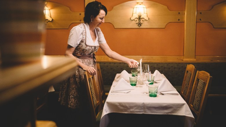 A woman in traditional dress lights a candle on a laid table in an inn.