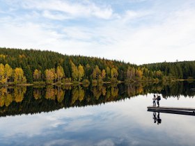 Die sanften H&uuml;gel spiegeln sich im ruhigen Wasser des Schlesinger Teichs, w&auml;hrend die bunten Bl&auml;tter der B&auml;ume den Herbst in voller Pracht zeigen. Ein romantischer Moment f&uuml;r Paare, die die friedliche Natur und die frische Luft genie&szlig;en m&ouml;chten.