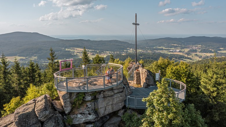 Aussichtsplattform auf einem Berg mit Kreuz und Blick auf bewaldete Landschaft.