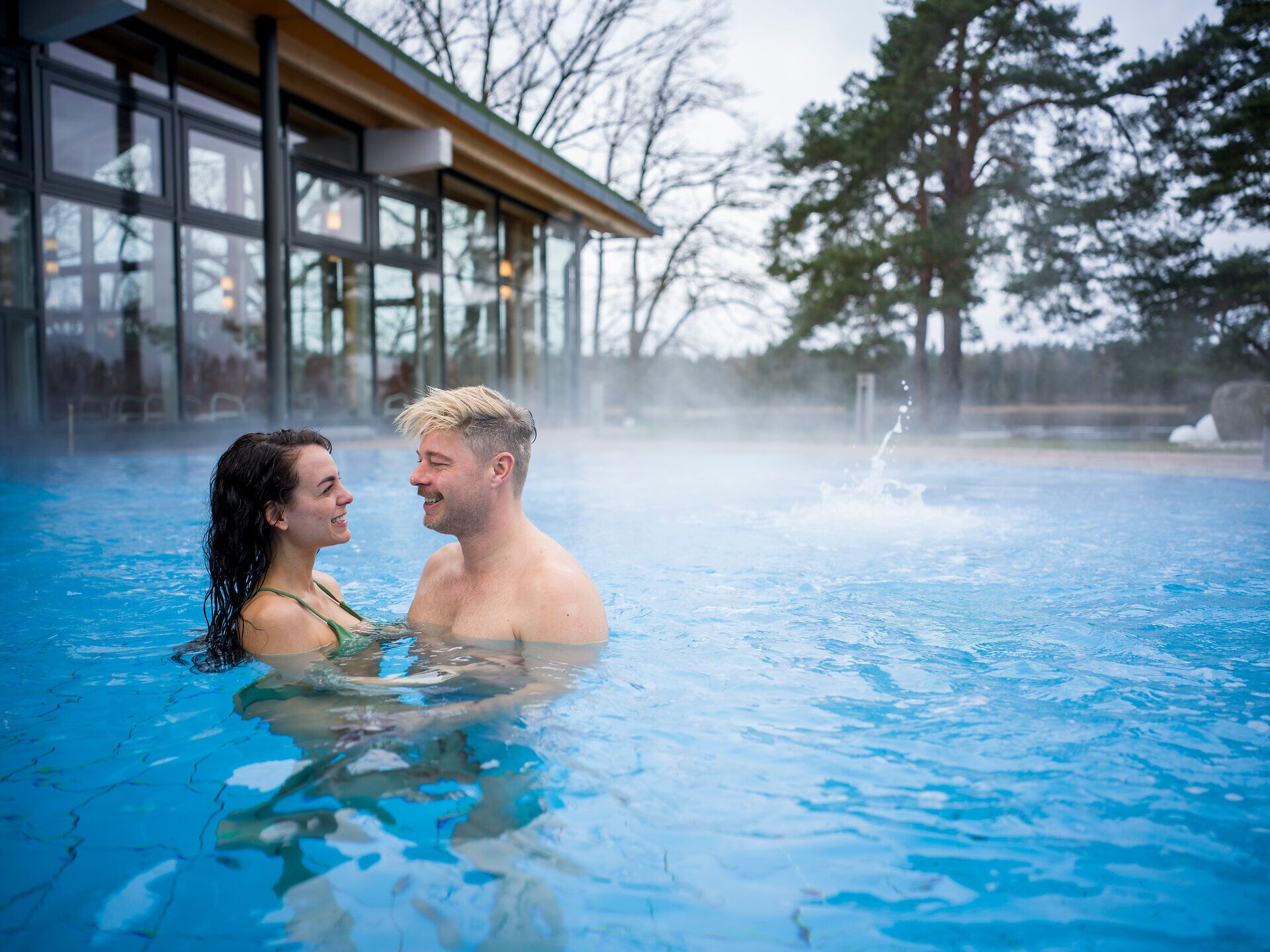 In einem einladenden Außenbecken genießen zwei Menschen die wohltuende Wärme des Thermalwassers, umgeben von einer malerischen Landschaft. Der Dampf, der sanft über der Wasseroberfläche schwebt, schafft eine romantische Atmosphäre, die zum Entspannen einlädt.