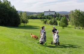 Zwei Golfer mit Hund auf gr&uuml;nem Golfplatz, Schloss im Hintergrund.
