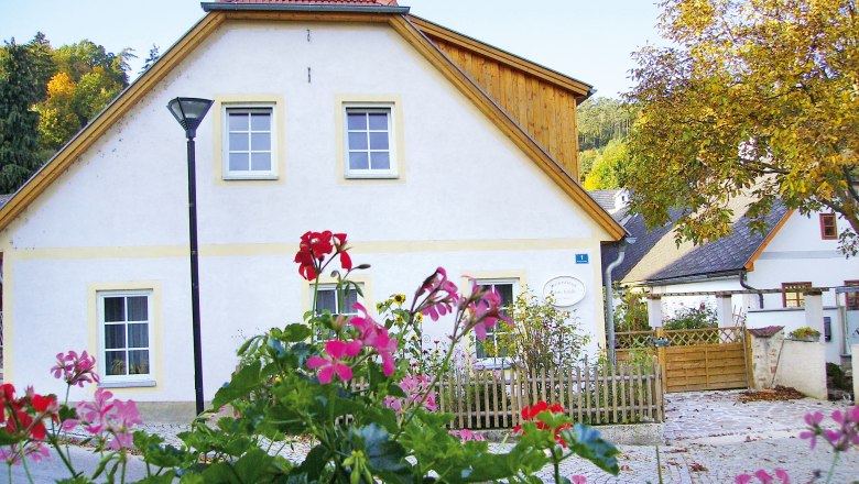 Ein weißes Ferienhaus mit roten Dachziegeln und einem Gartenzaun, umgeben von blühenden Blumen und Bäumen im Herbst.