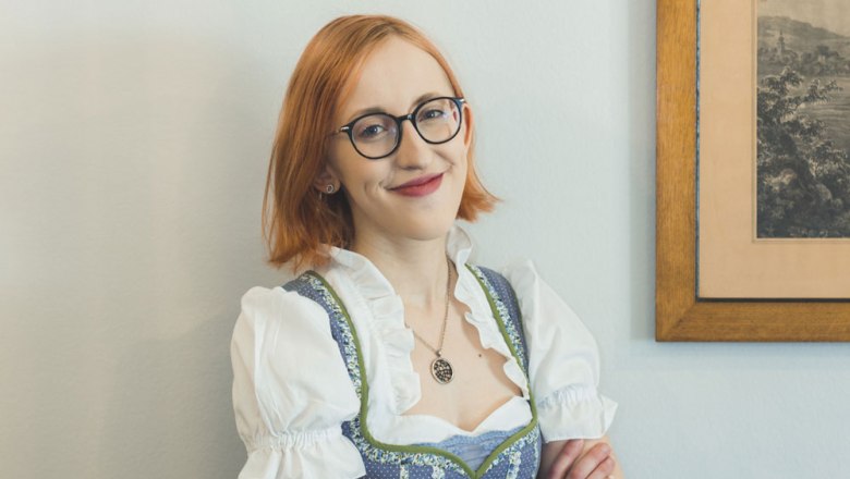 Eine Frau mit Brille und Dirndl lächelt vor einem Bild an der Wand.