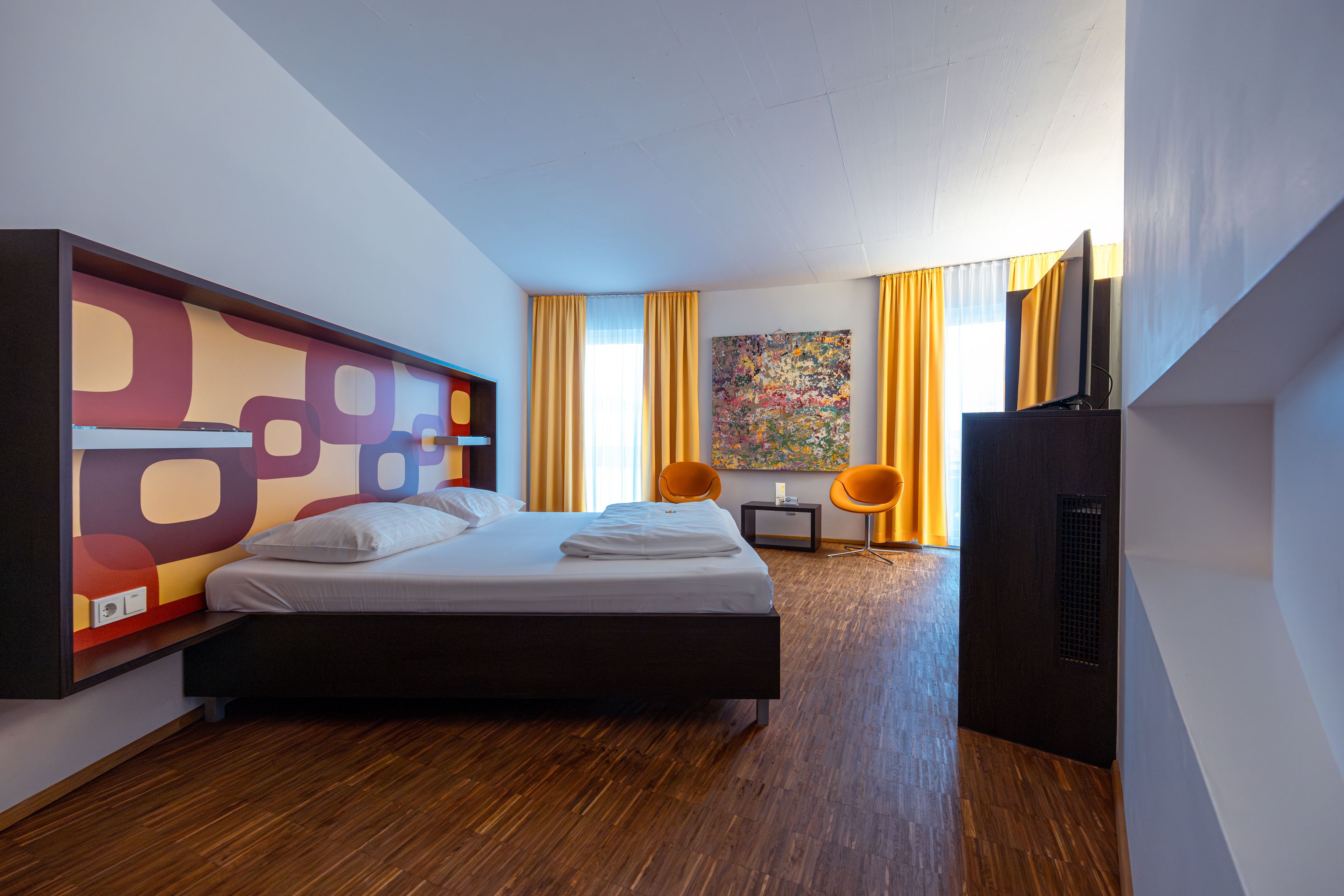 Modernes Hotelzimmer mit Doppelbett, buntem Wandmuster, zwei orangefarbenen Stühlen und gelben Vorhängen.