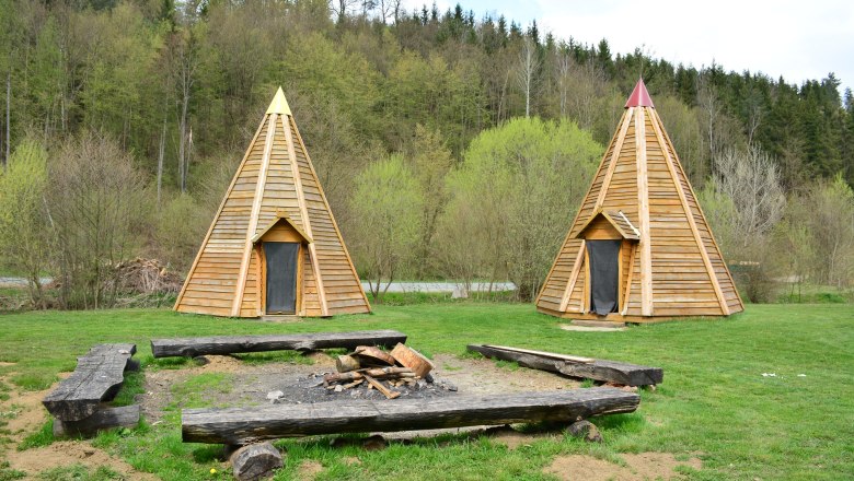 Two wooden tepees on a meadow in front of a forest.